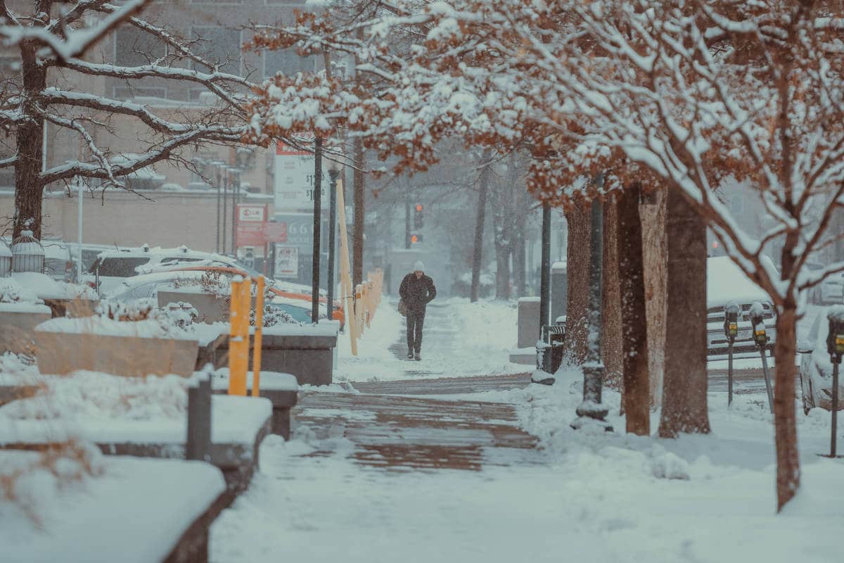 Person walking down snowy sidewalk passing by parking lots and buildings